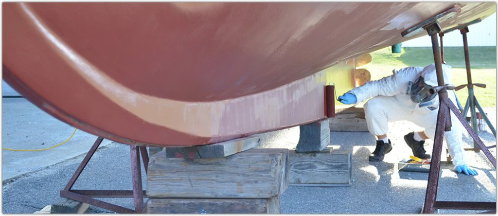 Person in protective personal equipment applying paint to the bottom of a boat hull