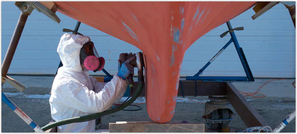 Person in paint suit and respirator sanding boat bottom to prepare for painting